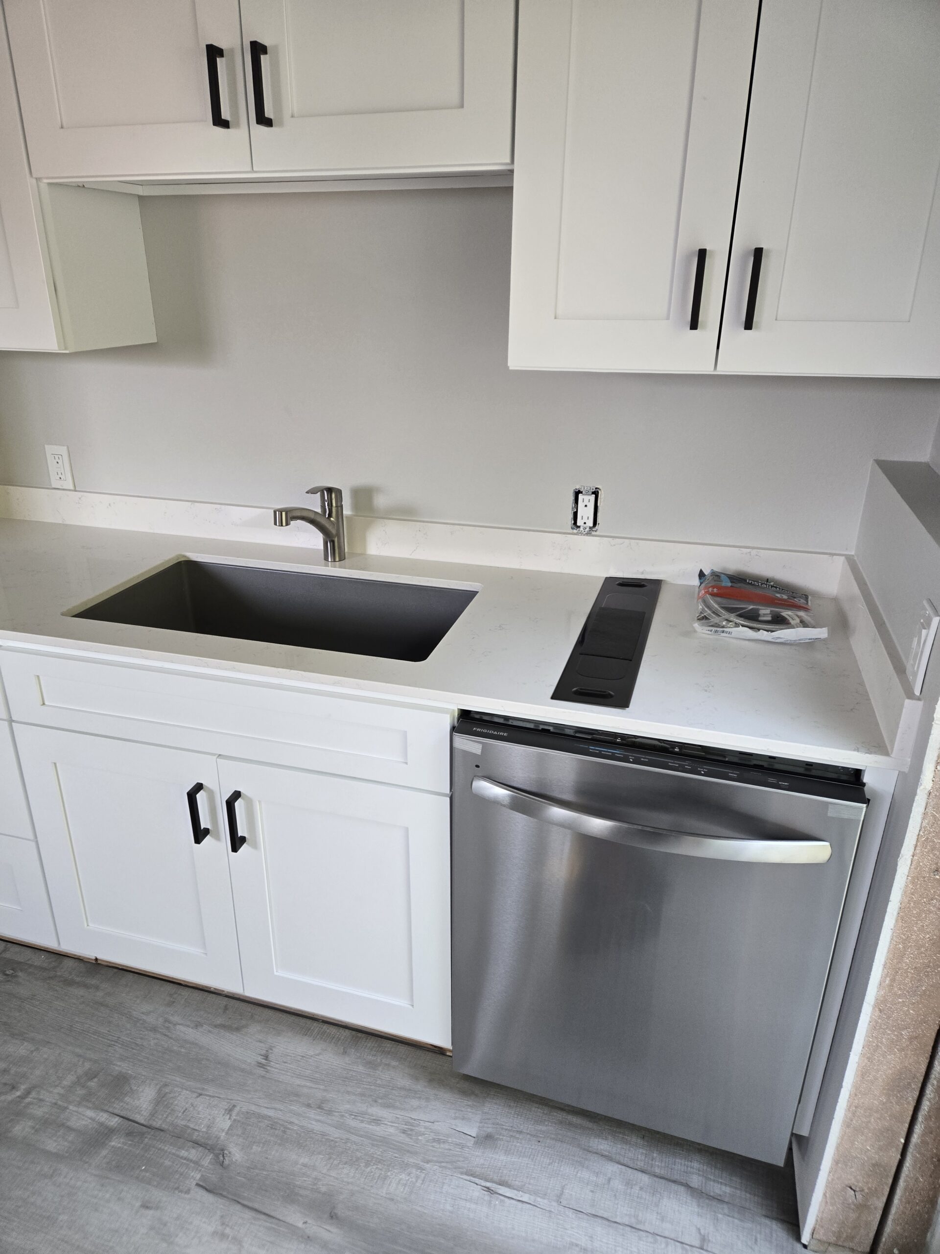 A modern kitchen with white cabinets, a stainless steel dishwasher, a single-basin sink, and an unfinished countertop with electrical outlets visible.