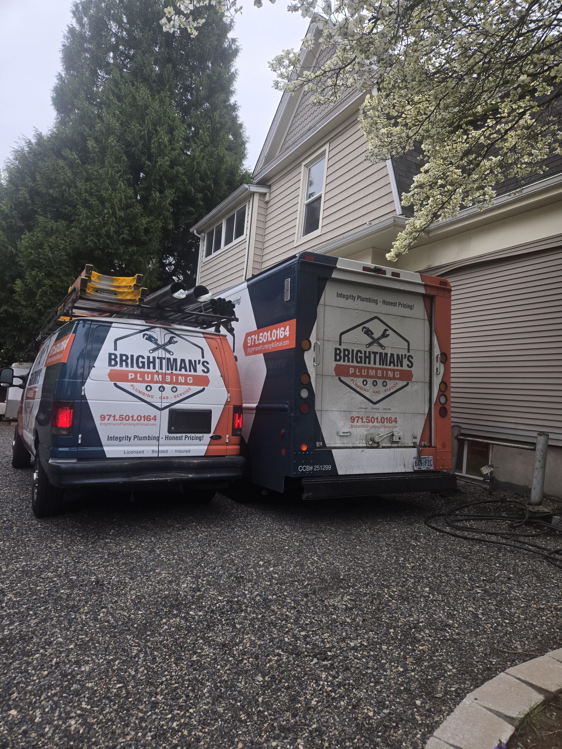 Two Brightman's Plumbing service vans are parked side by side in the driveway of a suburban house with beige siding and trees nearby.