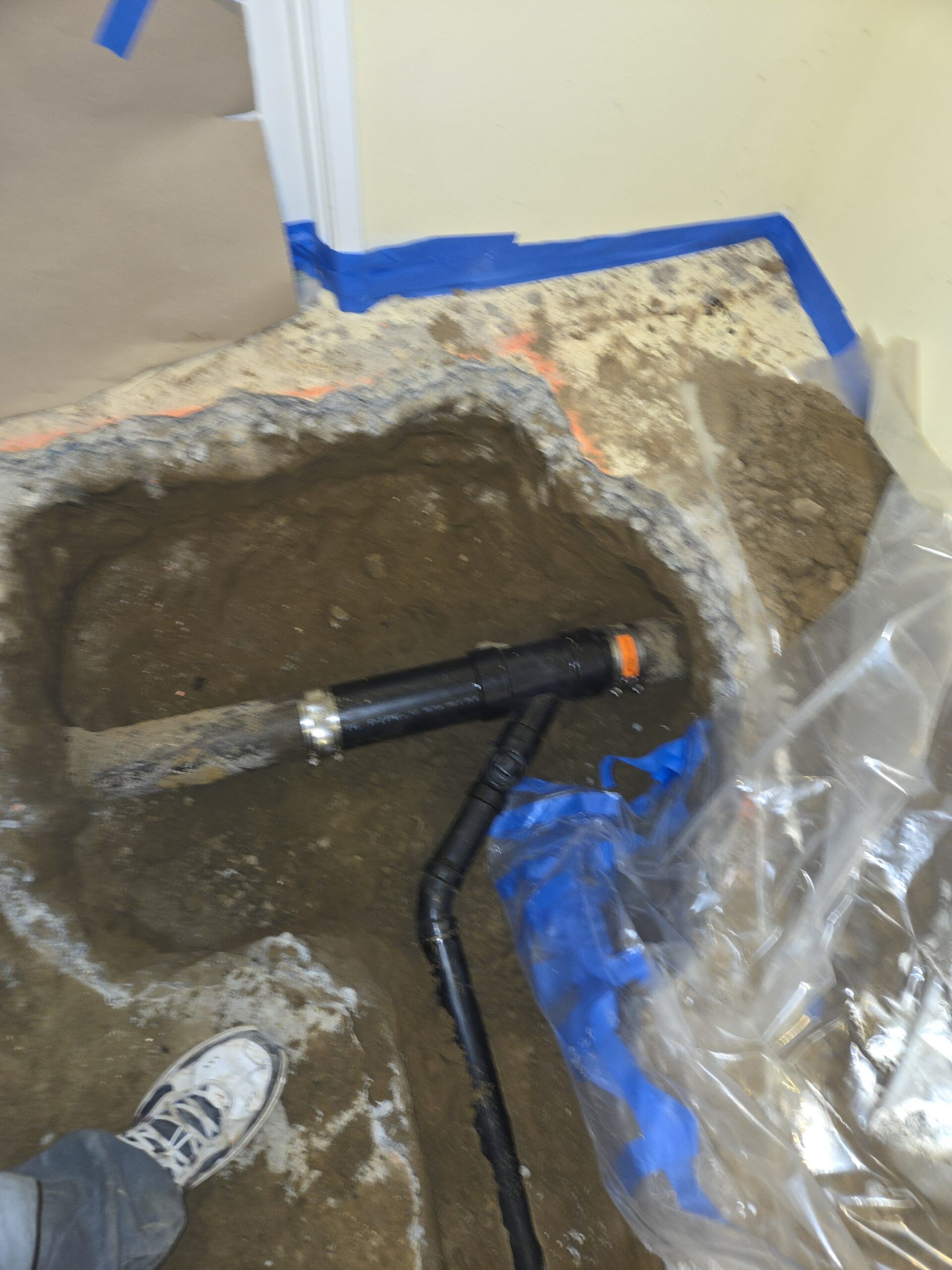 Large hole dug in a floor exposing soil and black pipes; a person's shoe is visible at the edge, with plastic sheeting and blue tape nearby.