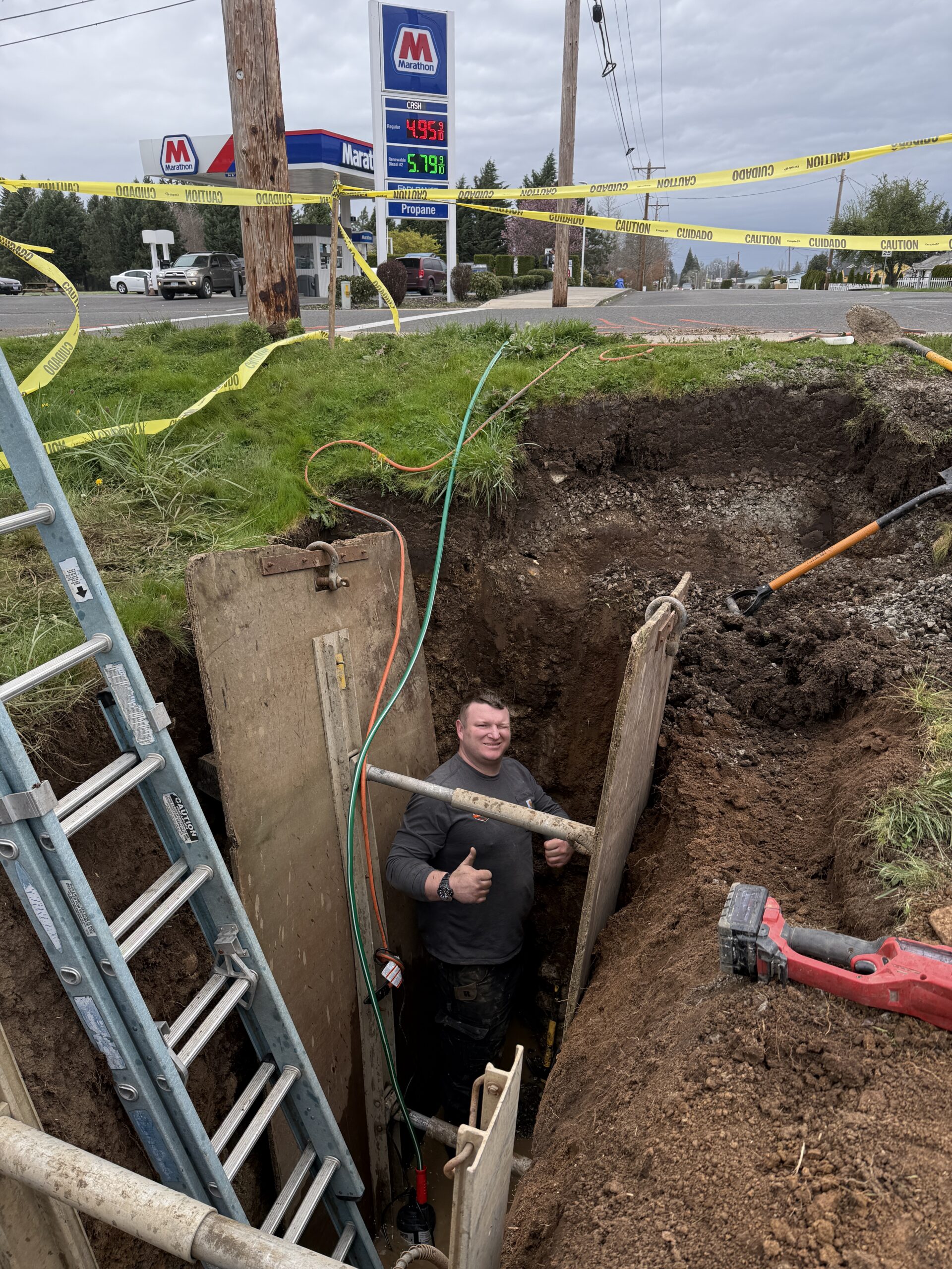A worker stands in a trench near a roadside, smiling and giving a thumbs-up. Construction equipment, caution tape, and a gas station are visible in the background.