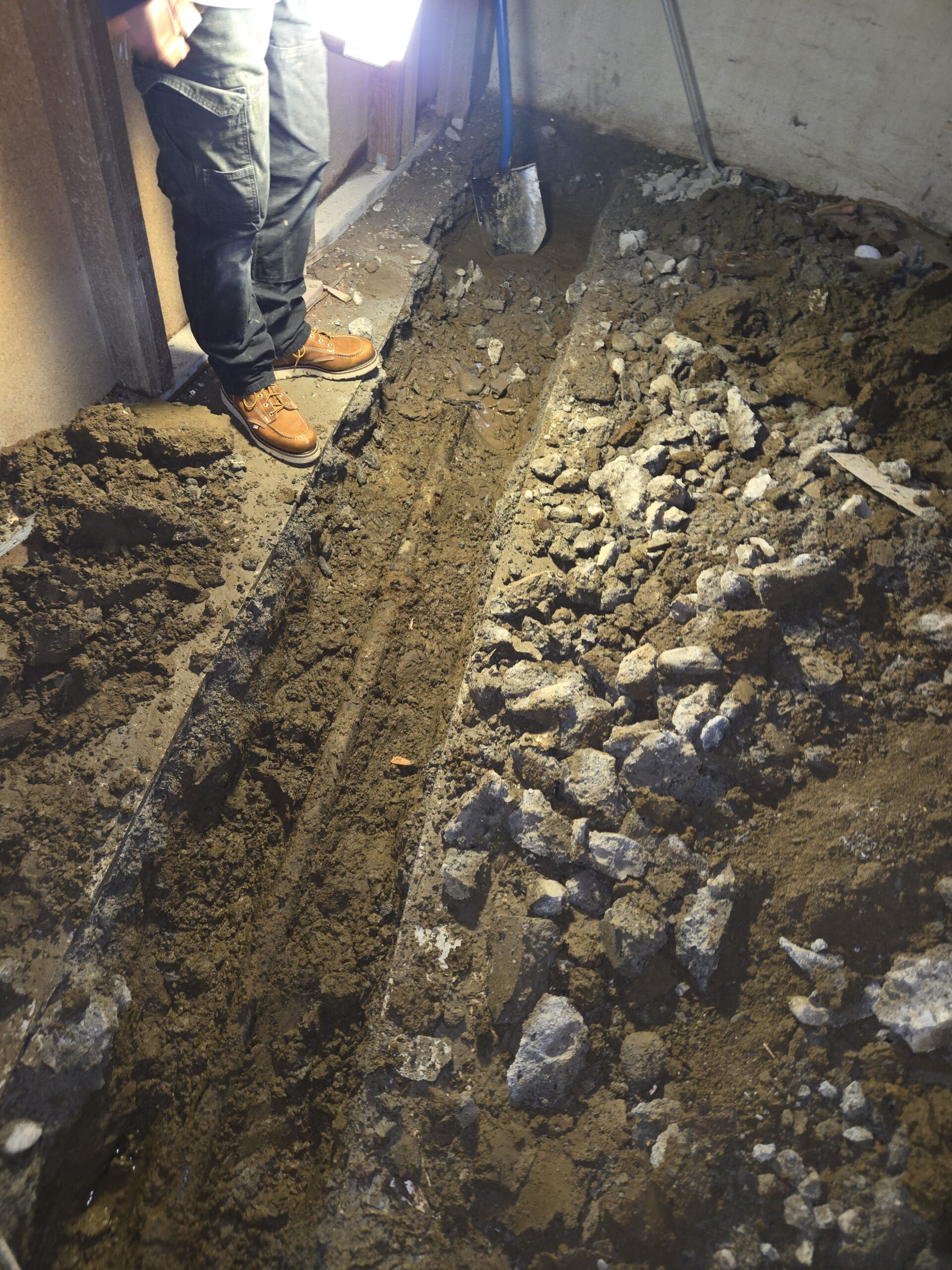 A person stands next to a narrow trench dug into the dirt and rubble floor of an indoor construction or renovation site.