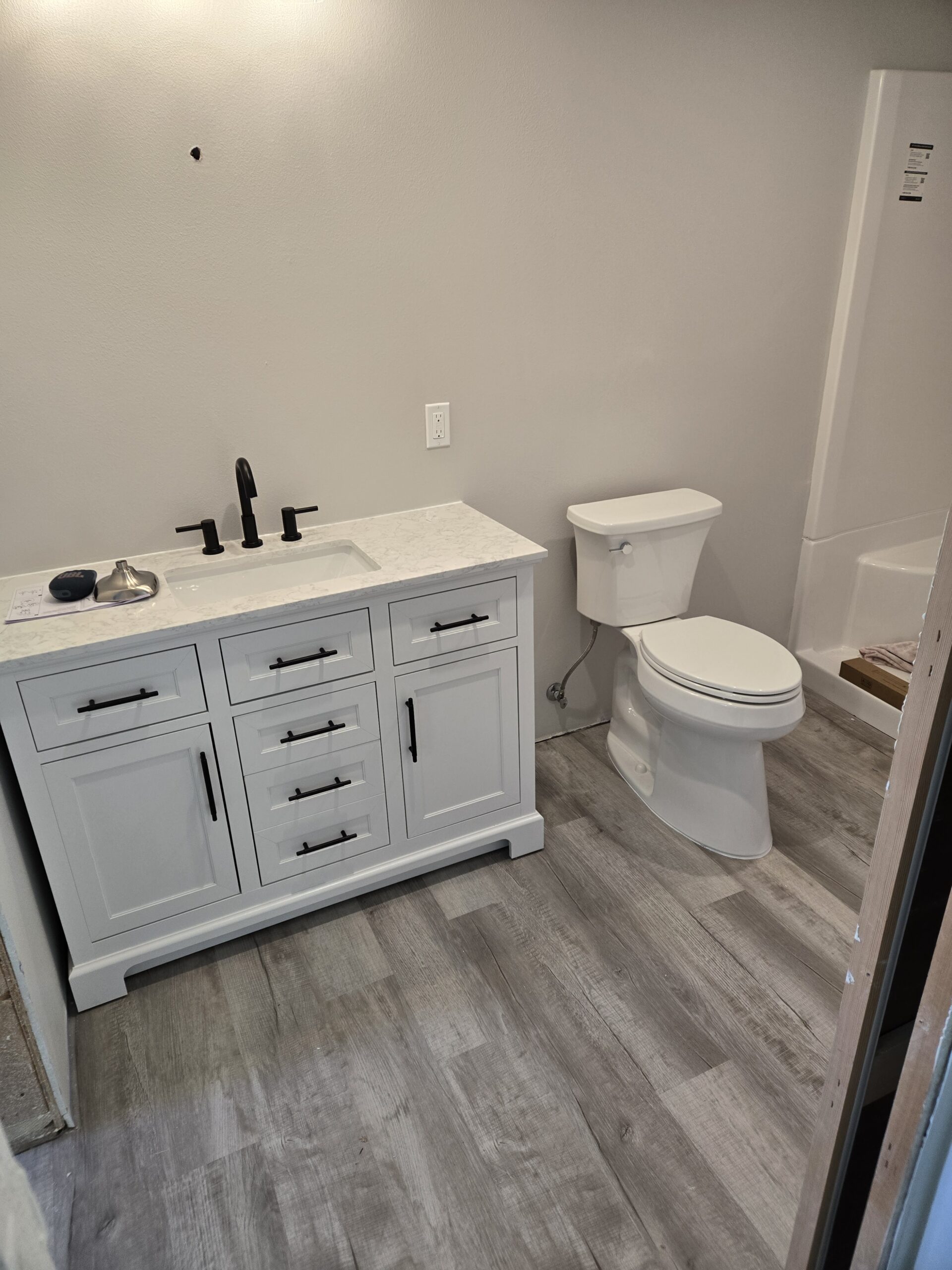 A bathroom with a white vanity and sink, black fixtures, a toilet, gray wood-look flooring, and a shower stall in the corner.