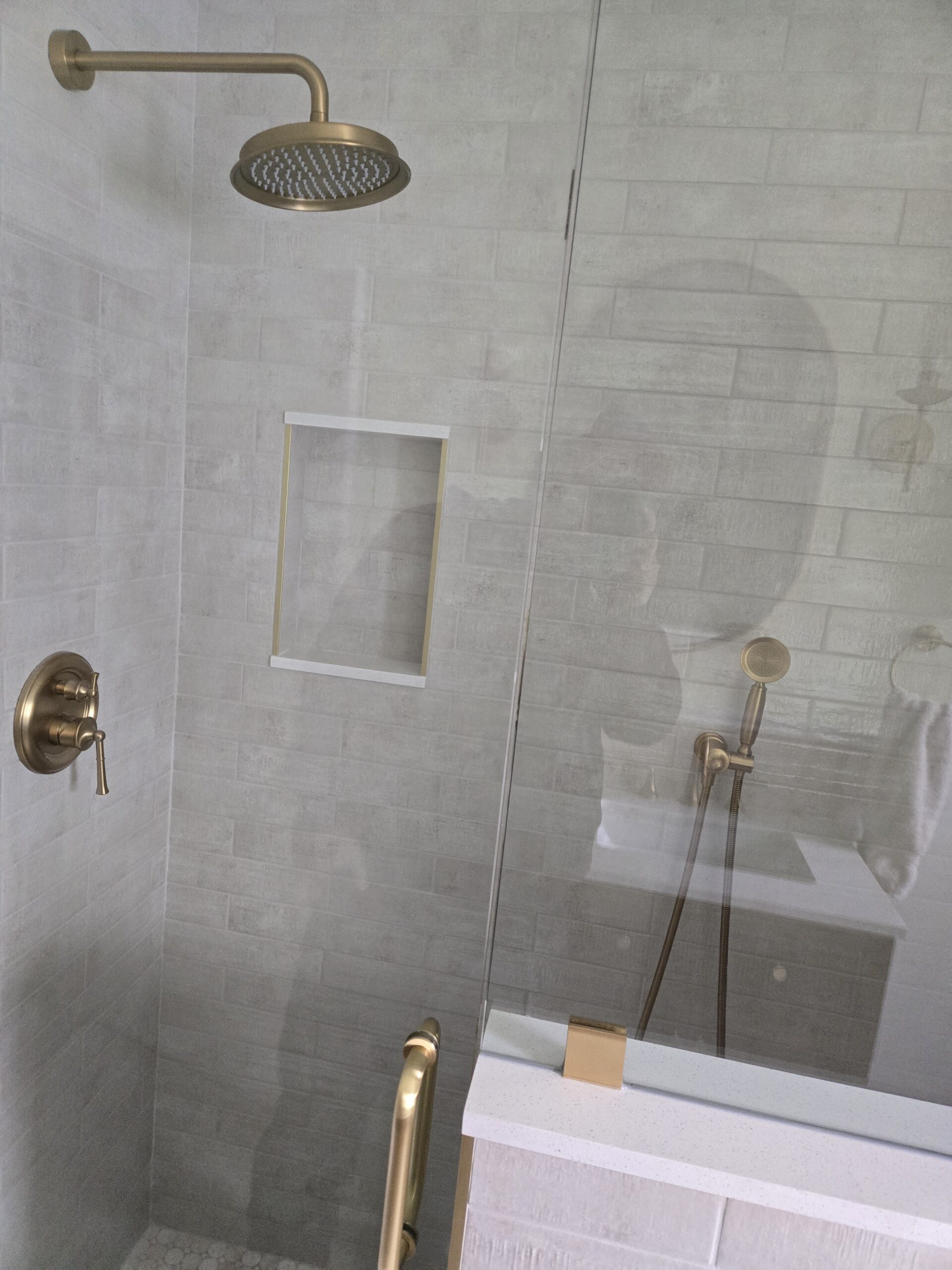 Modern bathroom shower with gold fixtures, including a rainfall showerhead, wall-mounted control, and a handheld shower next to a partial glass enclosure.