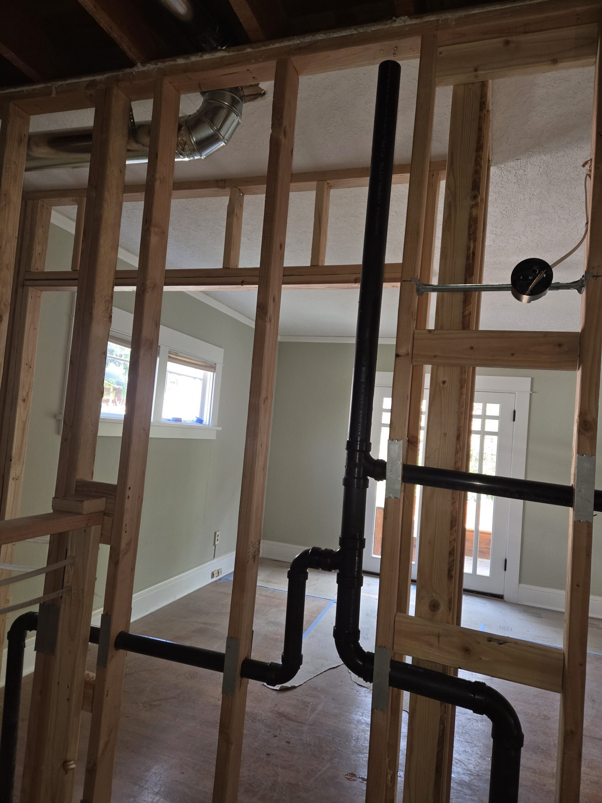 Exposed wall framing with black plumbing pipes and electrical wiring in a room under construction or renovation, with finished walls and windows visible in the background.