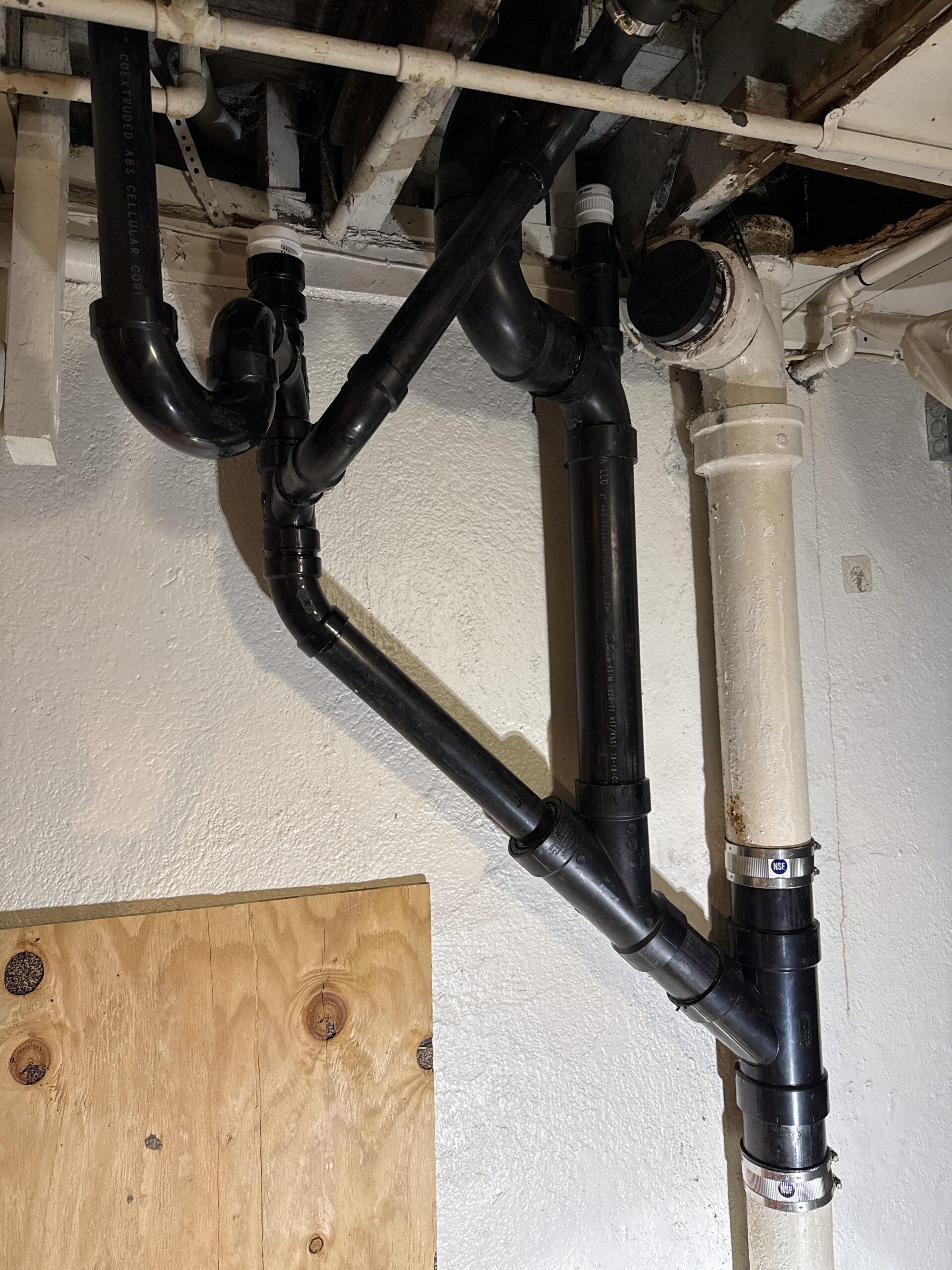 Several black PVC drainage pipes connected to a white vertical pipe on a basement wall, with visible joints and support brackets, near a plywood-covered opening.