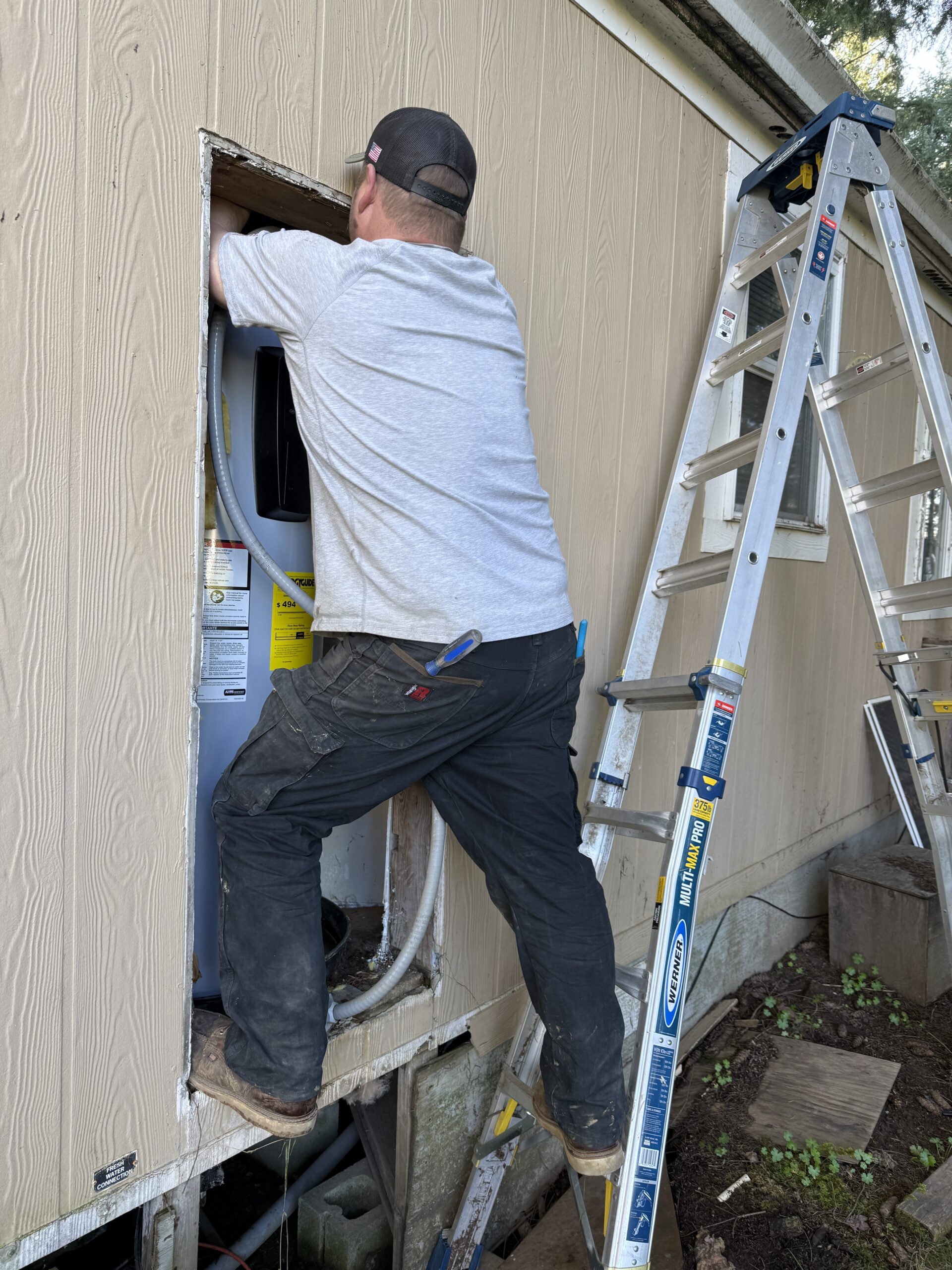A person stands on a ladder and works on installing or repairing an appliance through an opening in the exterior wall of a building.