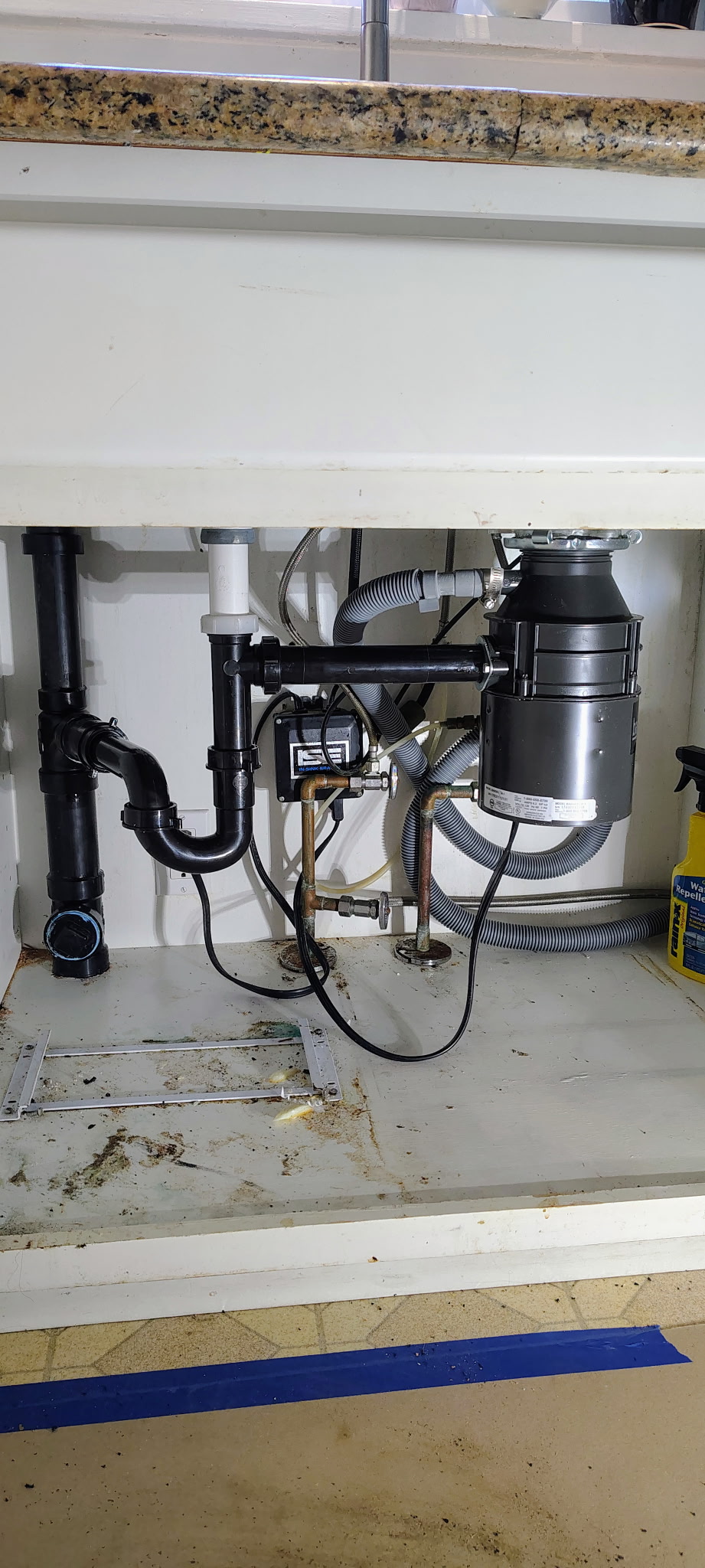 Plumbing and garbage disposal system installed under a kitchen sink, with visible pipes, hoses, and cleaning supplies.